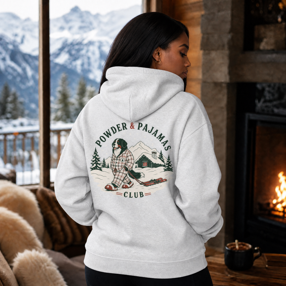 Person wearing a hoodie with 'Powder & Pajamas Club' design in a cozy cabin setting.