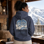 Person wearing a hoodie with a mountain and dog design, standing in front of a window with a snowy landscape.