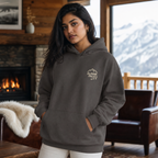 Woman wearing a dark heather gray hoodie with 'Alpine Pals' logo in a cozy living room with a fireplace and mountain view.