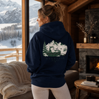Woman wearing a hoodie with a mountain design, looking out at snowy mountains from a cozy cabin.