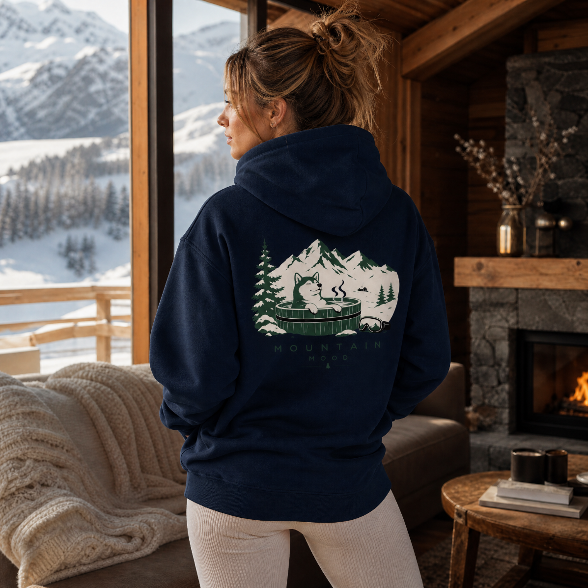 Woman wearing a hoodie with a mountain design, looking out at snowy mountains from a cozy cabin.