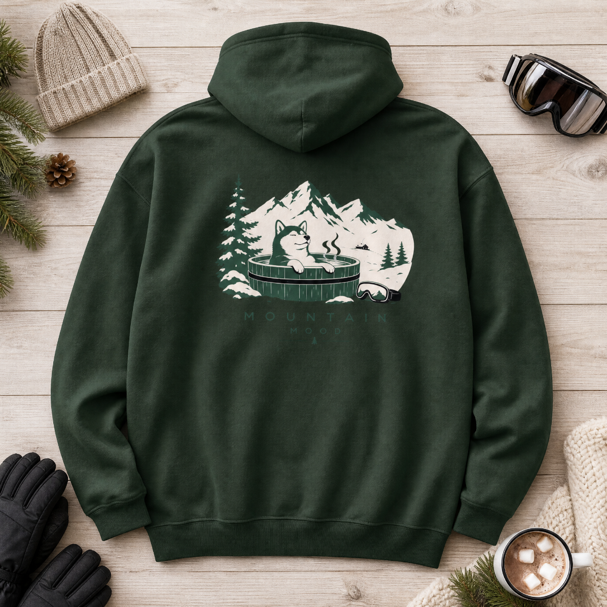 Forest green hoodie with mountain and bear design on a wooden surface with winter accessories.