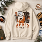 Sand colored hoodie with a graphic of a cat by a fireplace and 'APRES ALL DAY' text, surrounded by winter items on a wooden surface.