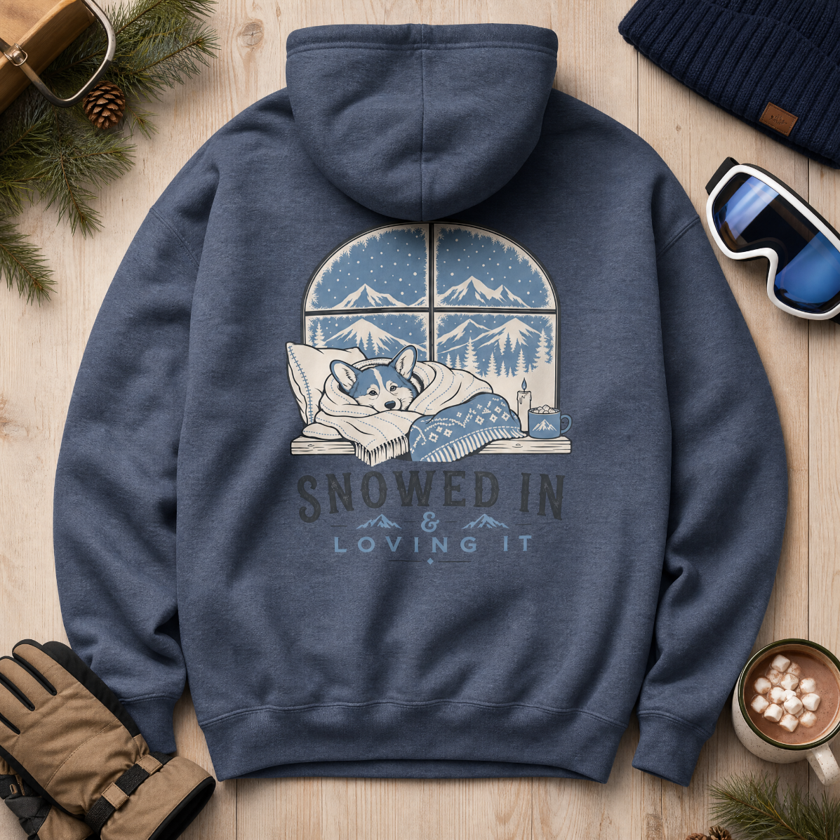 Heather sport dark navy hoodie with a graphic of a dog snuggled under a blanket, surrounded by snow, on a wooden surface with winter accessories.