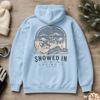 Light blue hoodie with a graphic of a person snuggling with a dog in front of a snowy window, text 'Snowed In & Loving It', on a wooden surface with winter decor.