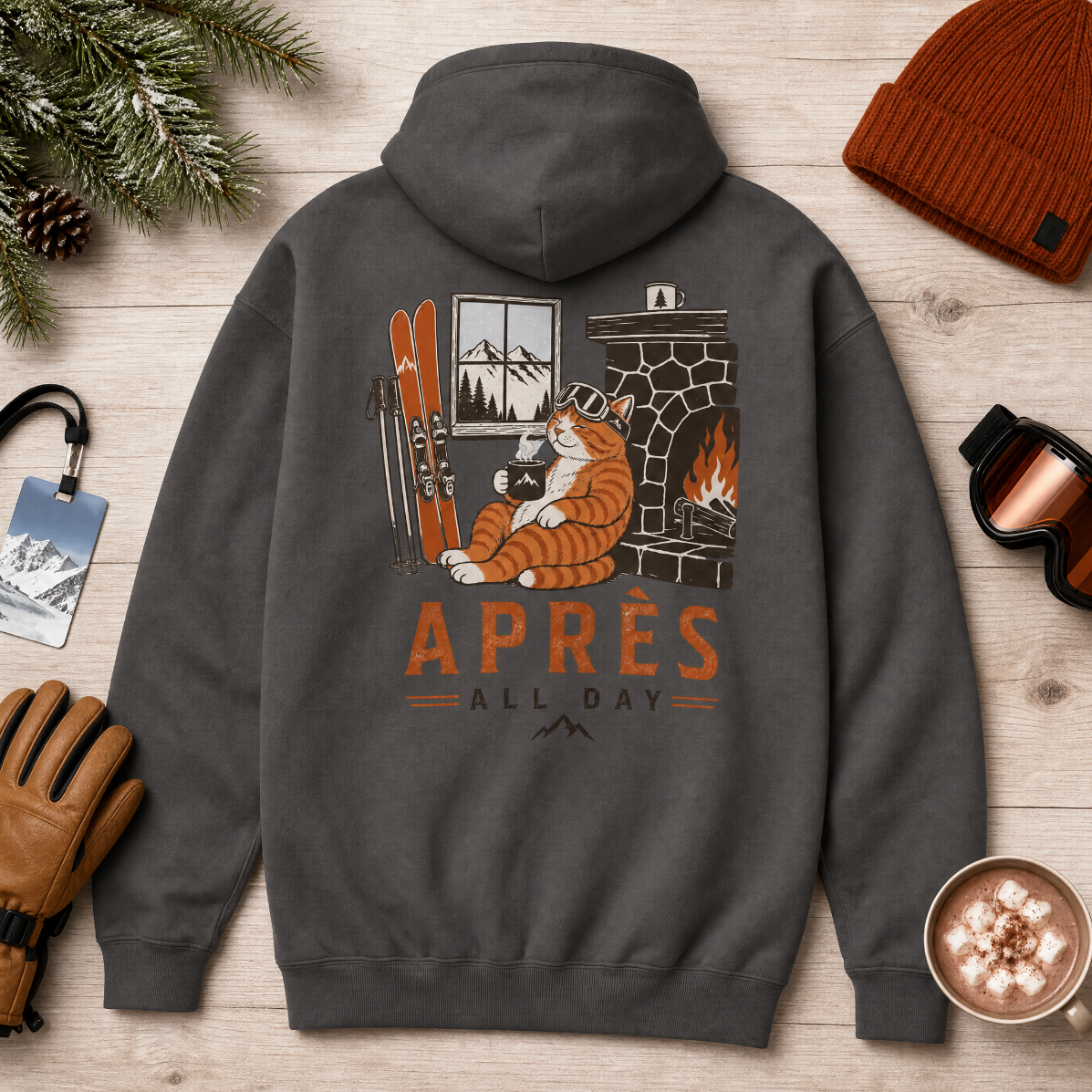Charcoal gray hoodie with a cat by a fireplace graphic and 'APRES ALL DAY' text on a wooden surface with winter accessories.