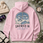 Pink hoodie with a winter-themed design and text 'Snowed In & Loving It' on a concrete surface with winter accessories.