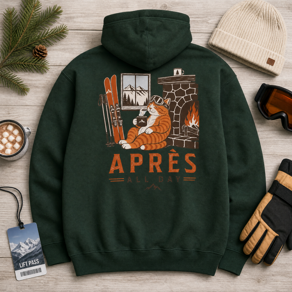 Forest green hoodie with a graphic of a cat by a fireplace and 'APRES ALL DAY' text, surrounded by winter accessories on a wooden surface.