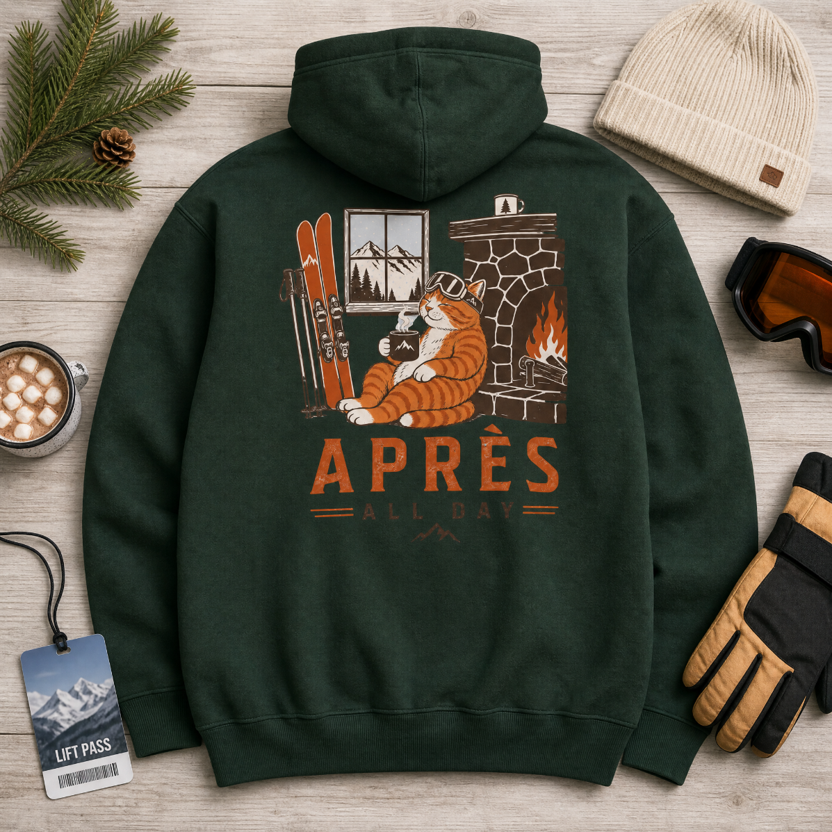 Forest green hoodie with a graphic of a cat by a fireplace and 'APRES ALL DAY' text, surrounded by winter accessories on a wooden surface.