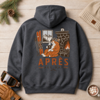Dark heather gray hoodie with a graphic of a cat by a fireplace and 'APRES ALL DAY' text, placed on a wooden surface with winter accessories.