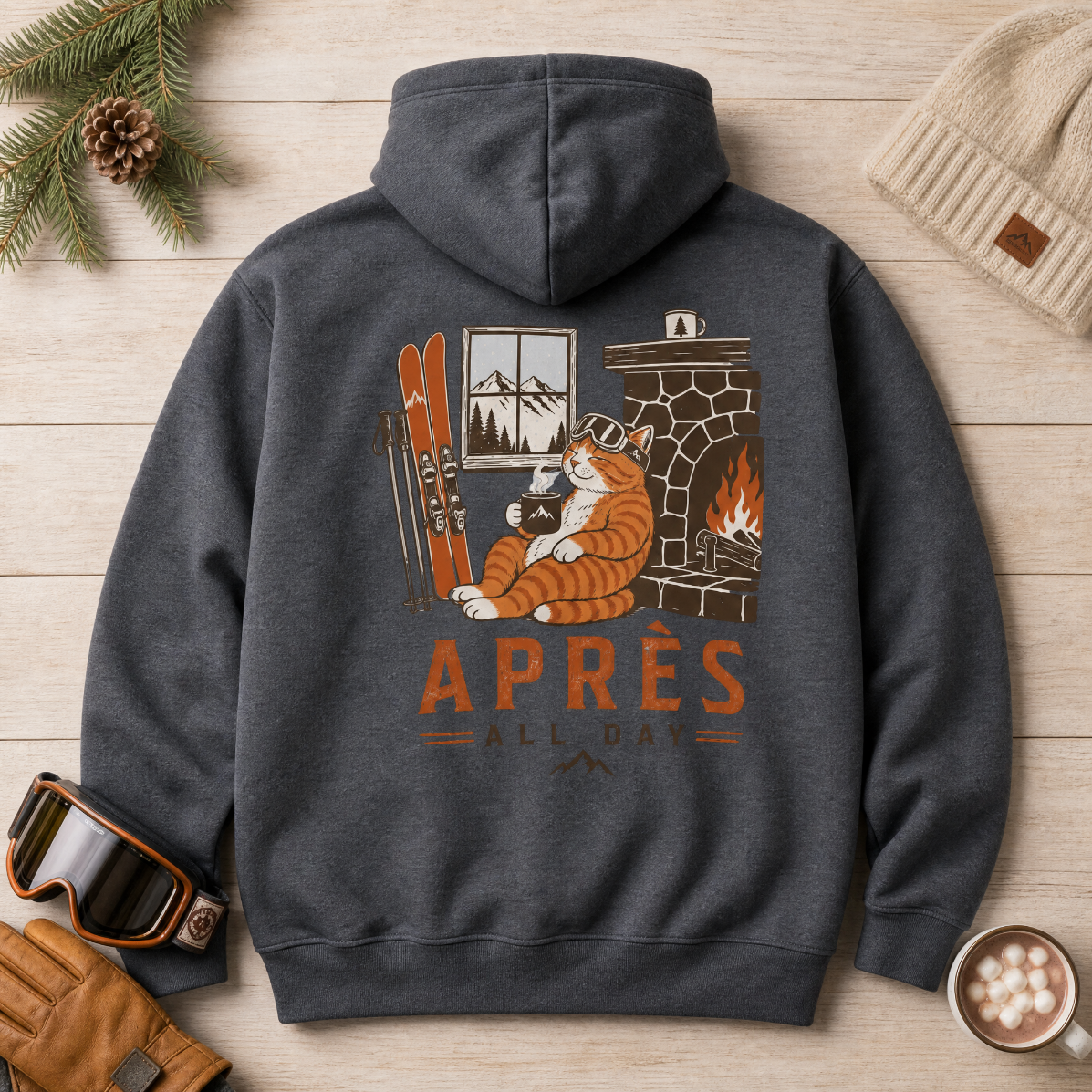 Dark heather gray hoodie with a graphic of a cat by a fireplace and 'APRES ALL DAY' text, placed on a wooden surface with winter accessories.