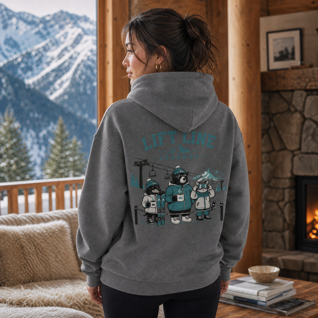 Person wearing a graphite gray hoodie with a mountain-themed design, standing in a cozy living room with a fireplace and snowy mountains outside.