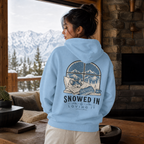 Person wearing a blue hoodie with a mountain and dog design, standing in a cozy room with a snowy landscape outside.