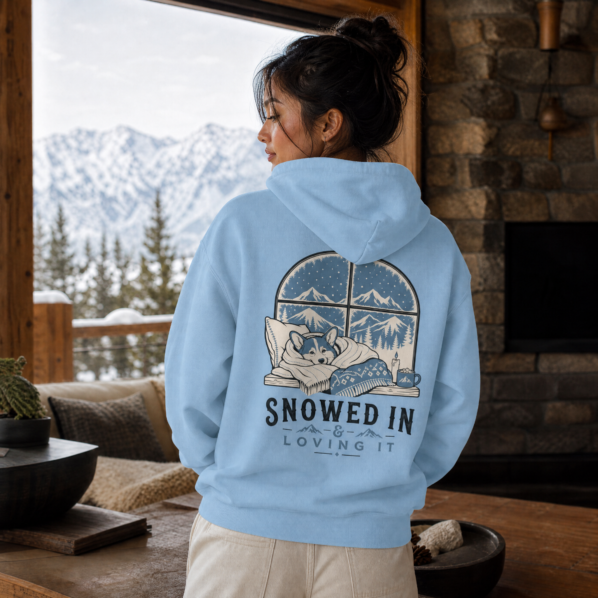 Person wearing a blue hoodie with a mountain and dog design, standing in a cozy room with a snowy landscape outside.