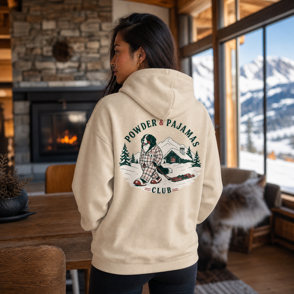 Woman wearing a sand colored hoodie that has a dog print with "POWDER & PAJAMAS CLUB" text.