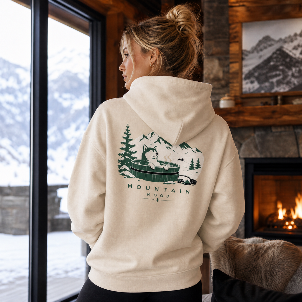 Person wearing a hoodie with a mountain and hot tub design, standing by a window with a snowy view.
