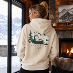 Person wearing a hoodie with a mountain and hot tub design, standing by a window with a snowy view.