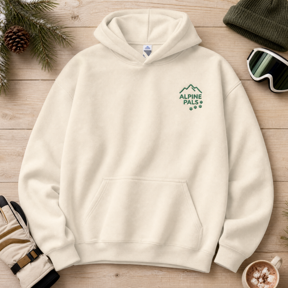 Sand colored hoodie with 'Alpine Pals' logo on a wooden surface with winter accessories.