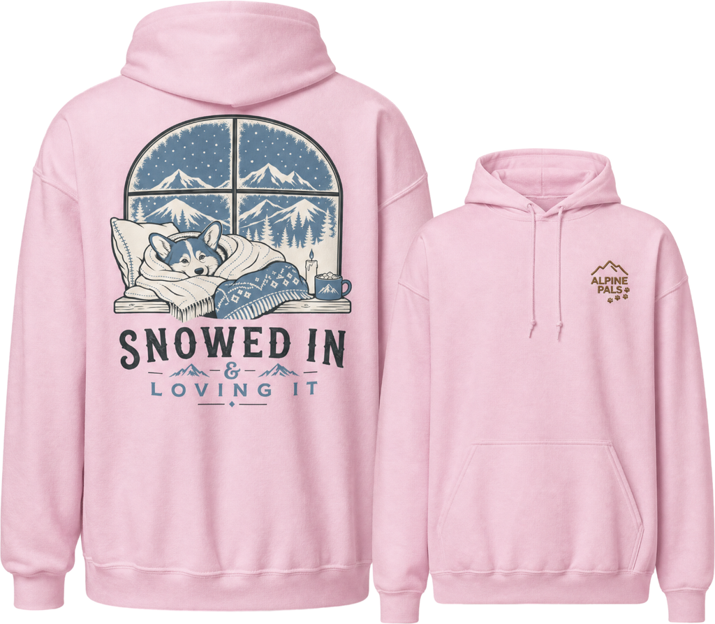 Pink hoodie with a graphic of a dog sleeping under a blanket and mountains, with text 'Snowed In & Loving It' on the front.
