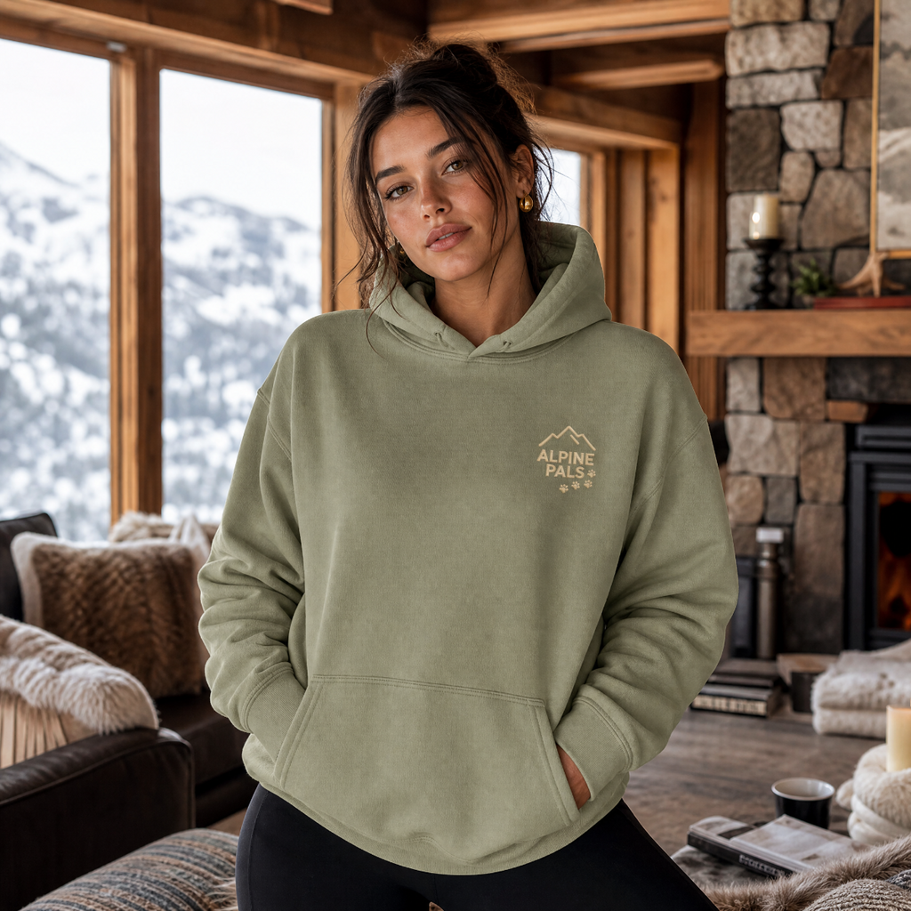 Woman wearing a military green hoodie with 'Alpine Pals' logo in a cozy indoor setting.