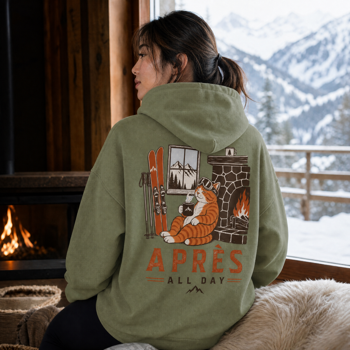 Person wearing a military green hoodie with a graphic design sitting by a fireplace with a snowy mountain view.