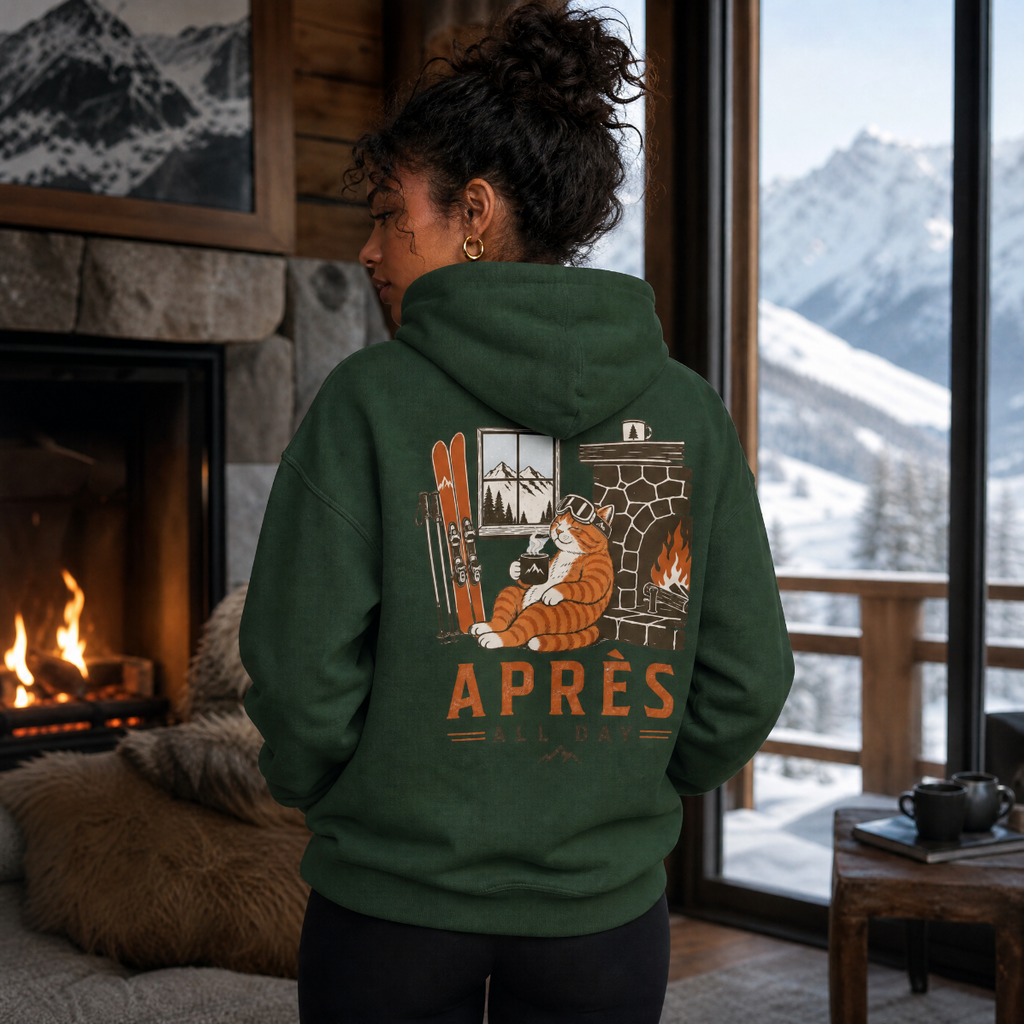 Person wearing a forest green hoodie with a mountain and cabin design, standing in a cozy room with a fireplace and snowy view.