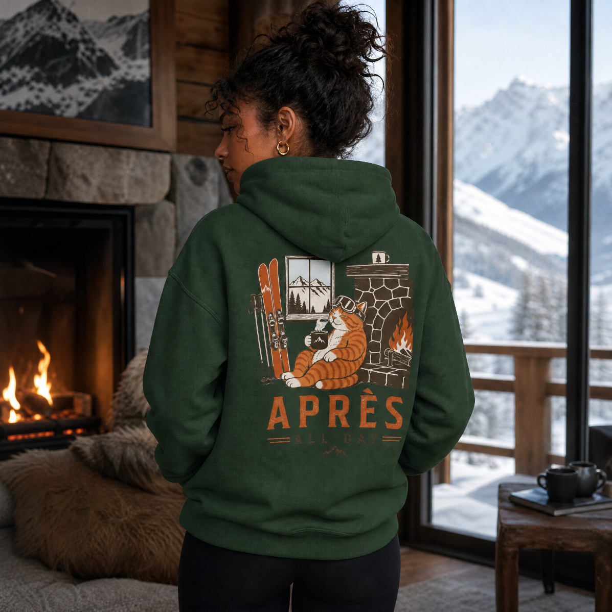 Person wearing a forest green hoodie with a mountain and cabin design, standing in a cozy room with a fireplace and snowy view.