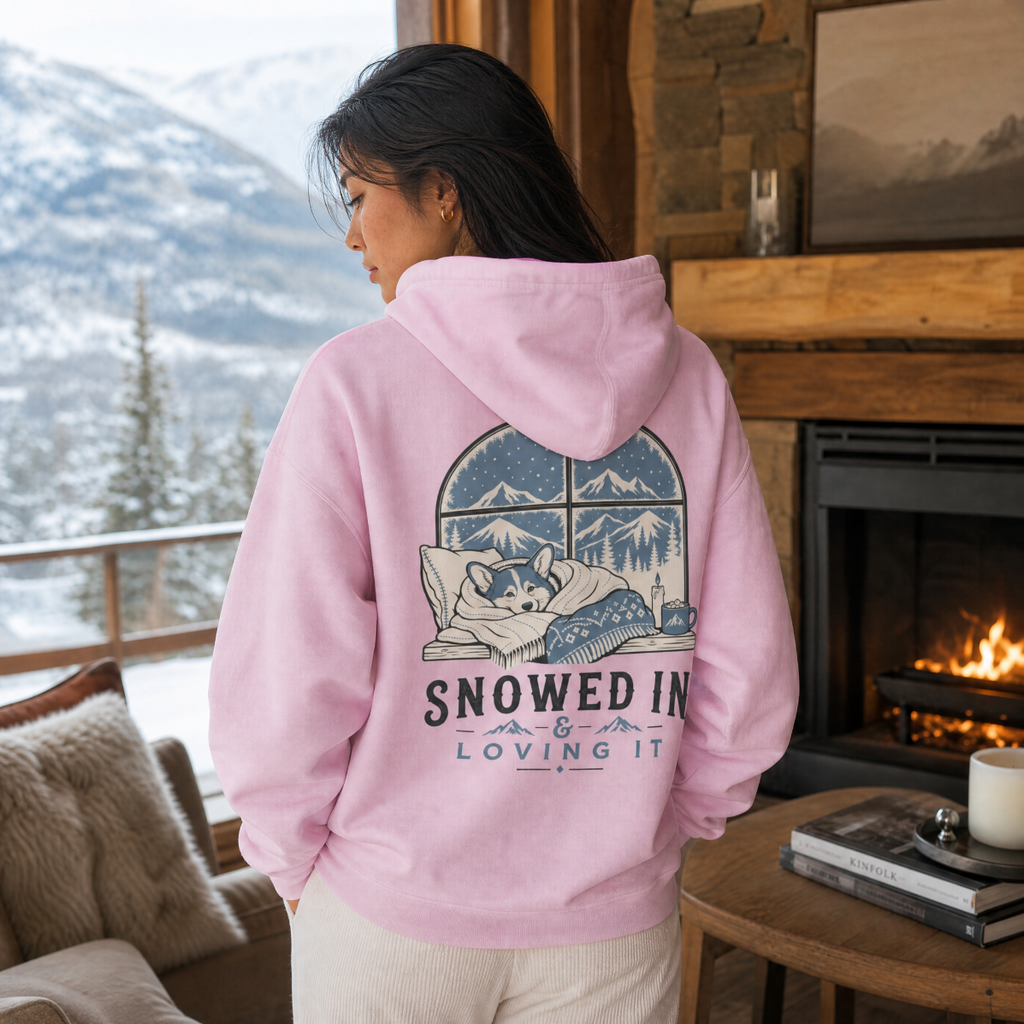 Person wearing a pink hoodie with a snowy design and text, standing in a cozy room with a fireplace and mountain view.