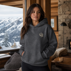 Woman wearing a dark heather gray hoodie with a logo, standing in a cozy room with a mountain view.