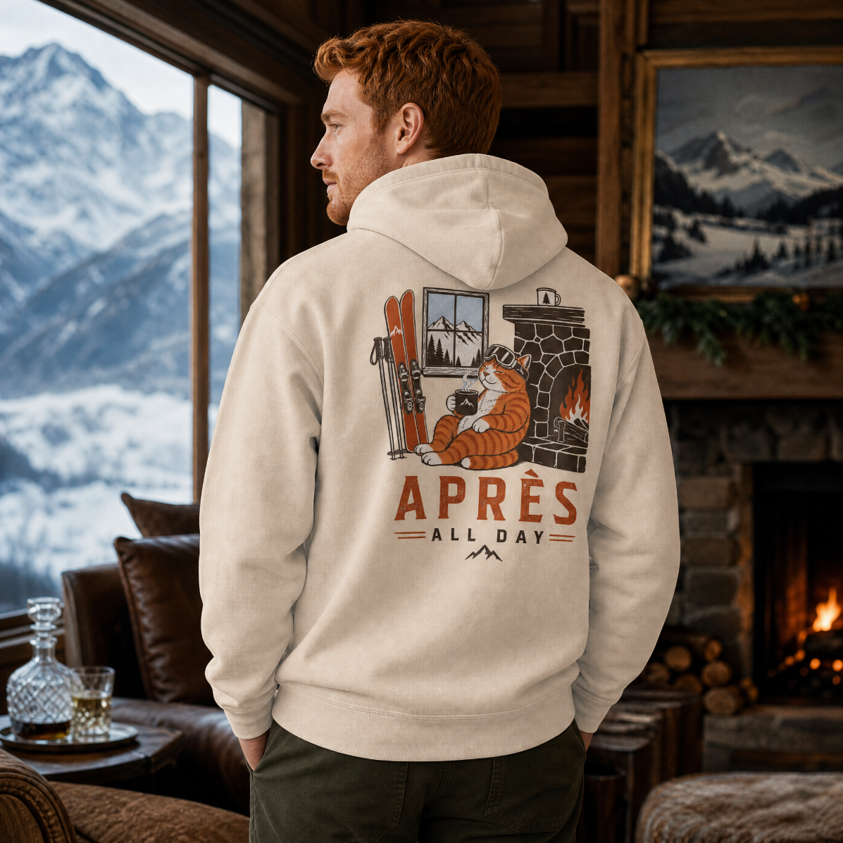 Man wearing a hoodie with a mountain and cabin design, standing in a cozy room with a fireplace and mountain view.