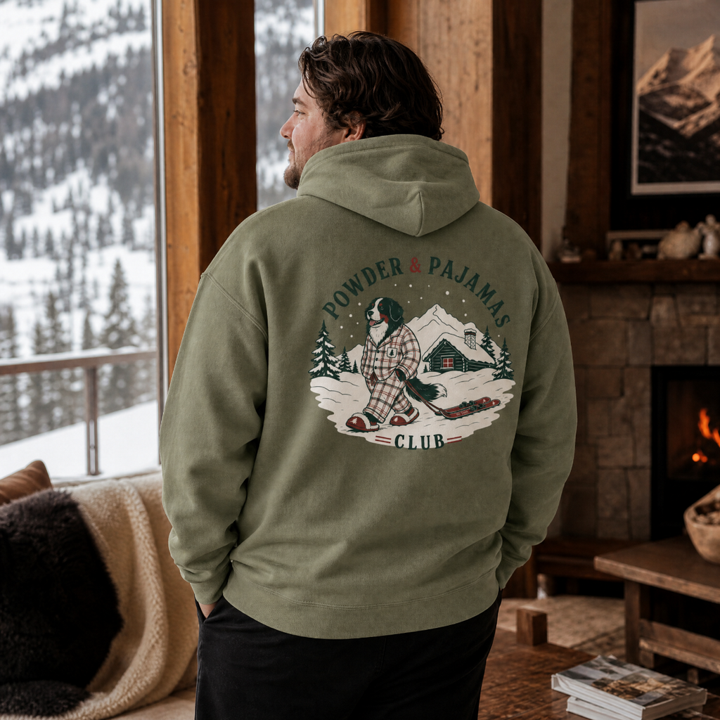 Man wearing a military green hoodie with a graphic design, standing in a cozy room with a mountain view.