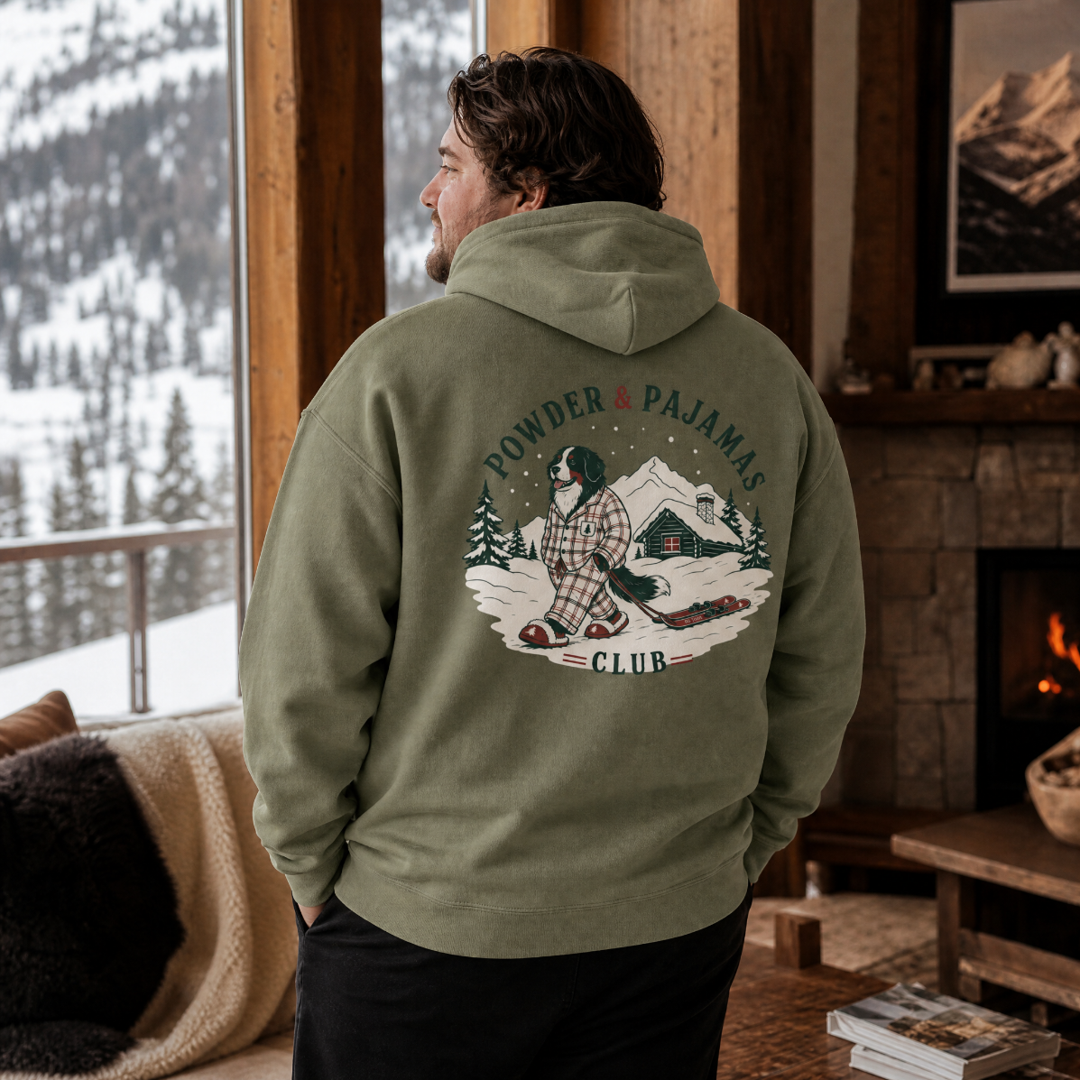 Man wearing a military green hoodie with a graphic design, standing in a cozy room with a mountain view.