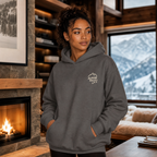 Woman wearing a charcoal gray hoodie with 'Alpine Pals' logo in a cozy living room with a fireplace and mountain view.