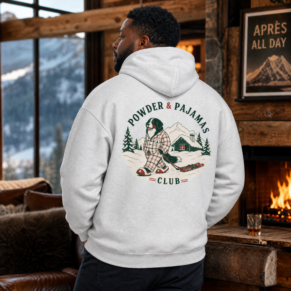 Person wearing a hoodie with a 'Powder & Pajamas Club' design in a cozy room with a fireplace and mountain view.