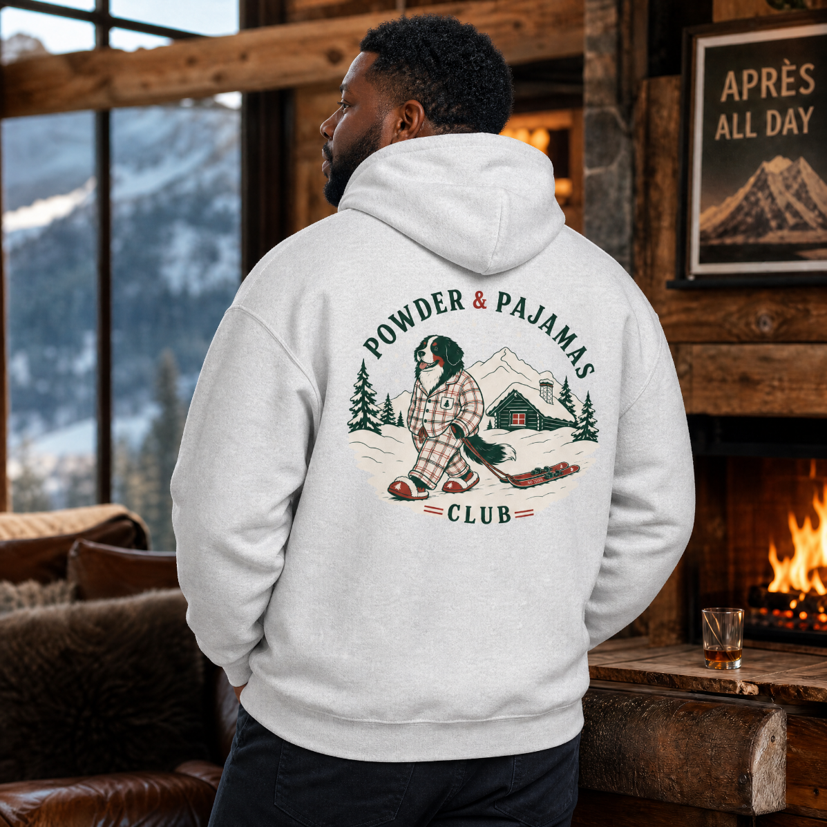 Person wearing a hoodie with a 'Powder & Pajamas Club' design in a cozy room with a fireplace and mountain view.