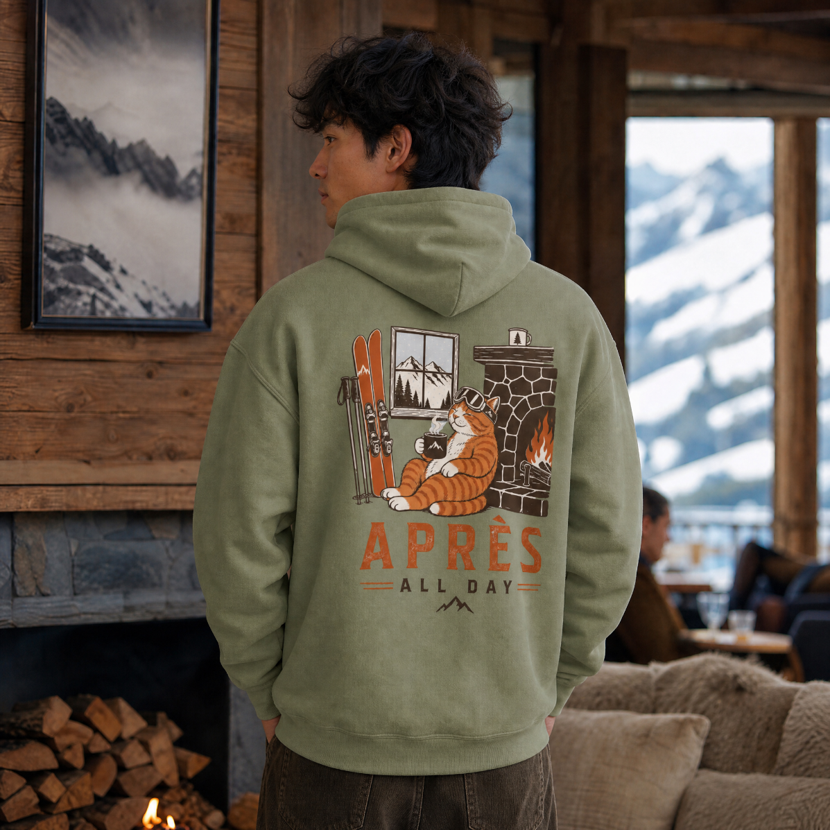 Person wearing a military green hoodie with a graphic design and text, standing in a cozy indoor setting with mountain views.
