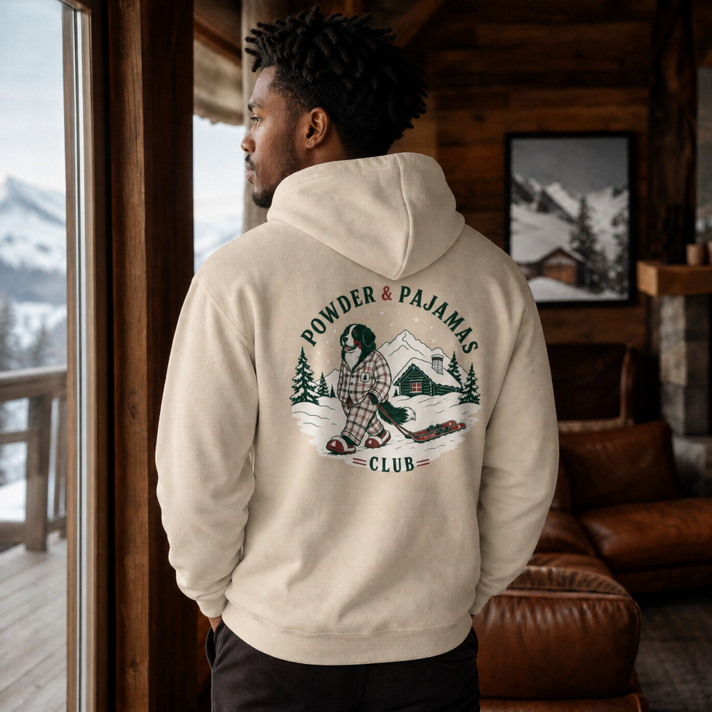 Man in a cozy chalet setting wearing a sand colored hoodie that has a dog print design with 'POWDER & PAJAMAS CLUB' text.