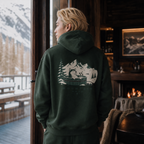 Person wearing a forest green hoodie with a mountain and cabin design, standing by a window with a snowy landscape view.