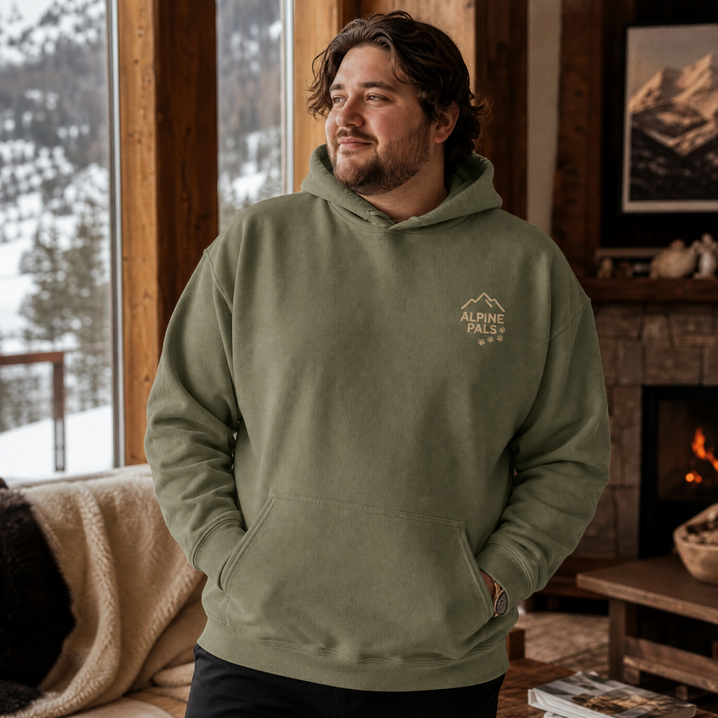 Man wearing a military green hoodie with a logo, standing in a cozy room with a fireplace and snowy outdoor view.