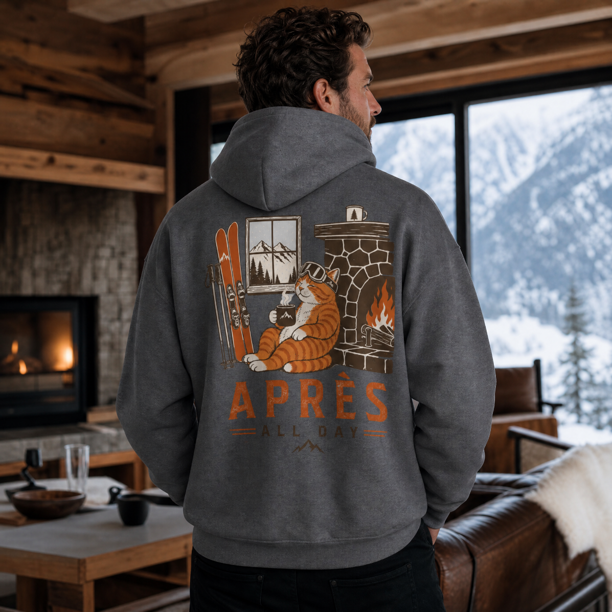 Man wearing a hoodie with a cat and fireplace design, standing in a cozy room with a mountain view.