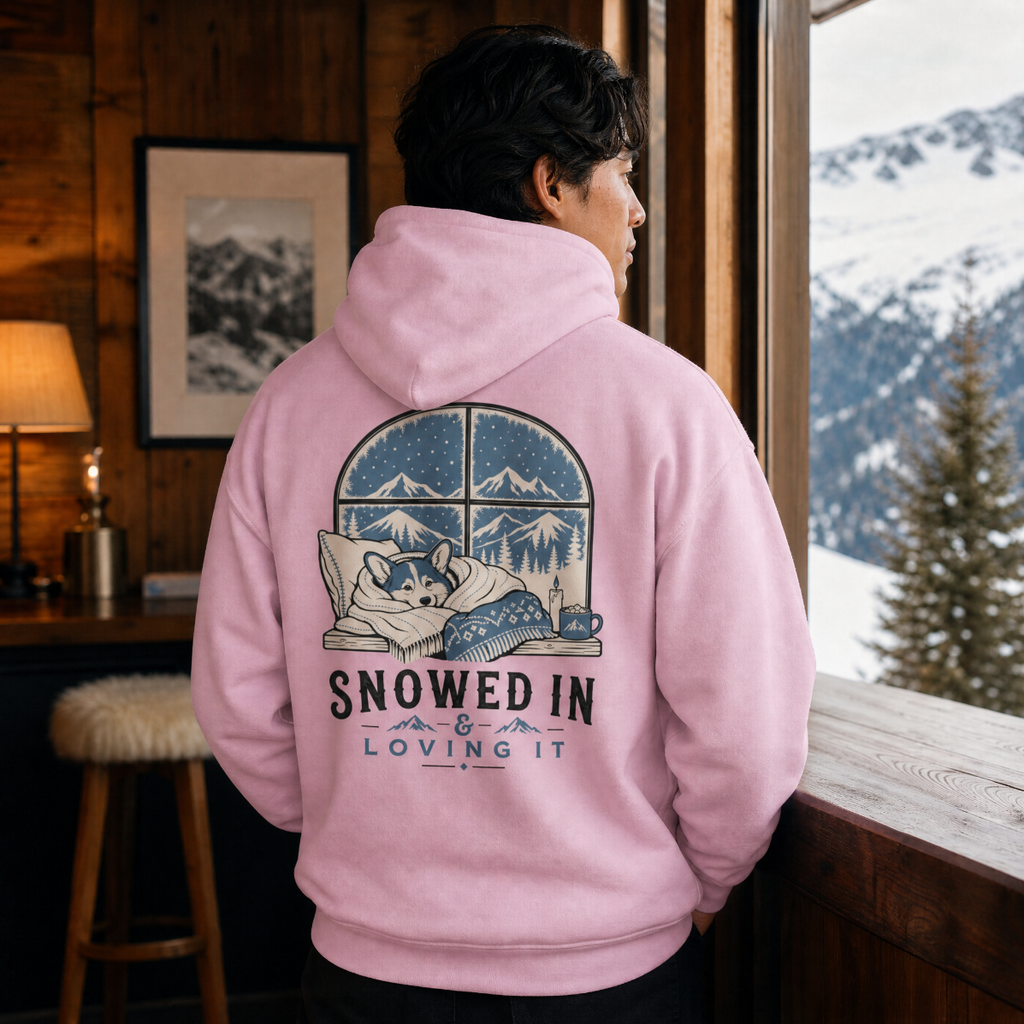 Person wearing a pink hoodie with a graphic of a dog and text 'Snowed In & Loving It' in a mountain cabin.