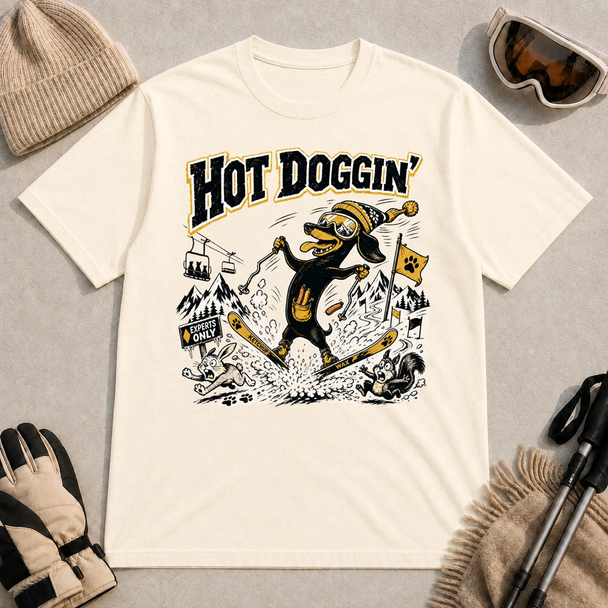 Natural tee featuring a goggle-wearing dog skiing in a spread-eagle pose while startling woodland animals, with the phrase “Hot Doggin’.”