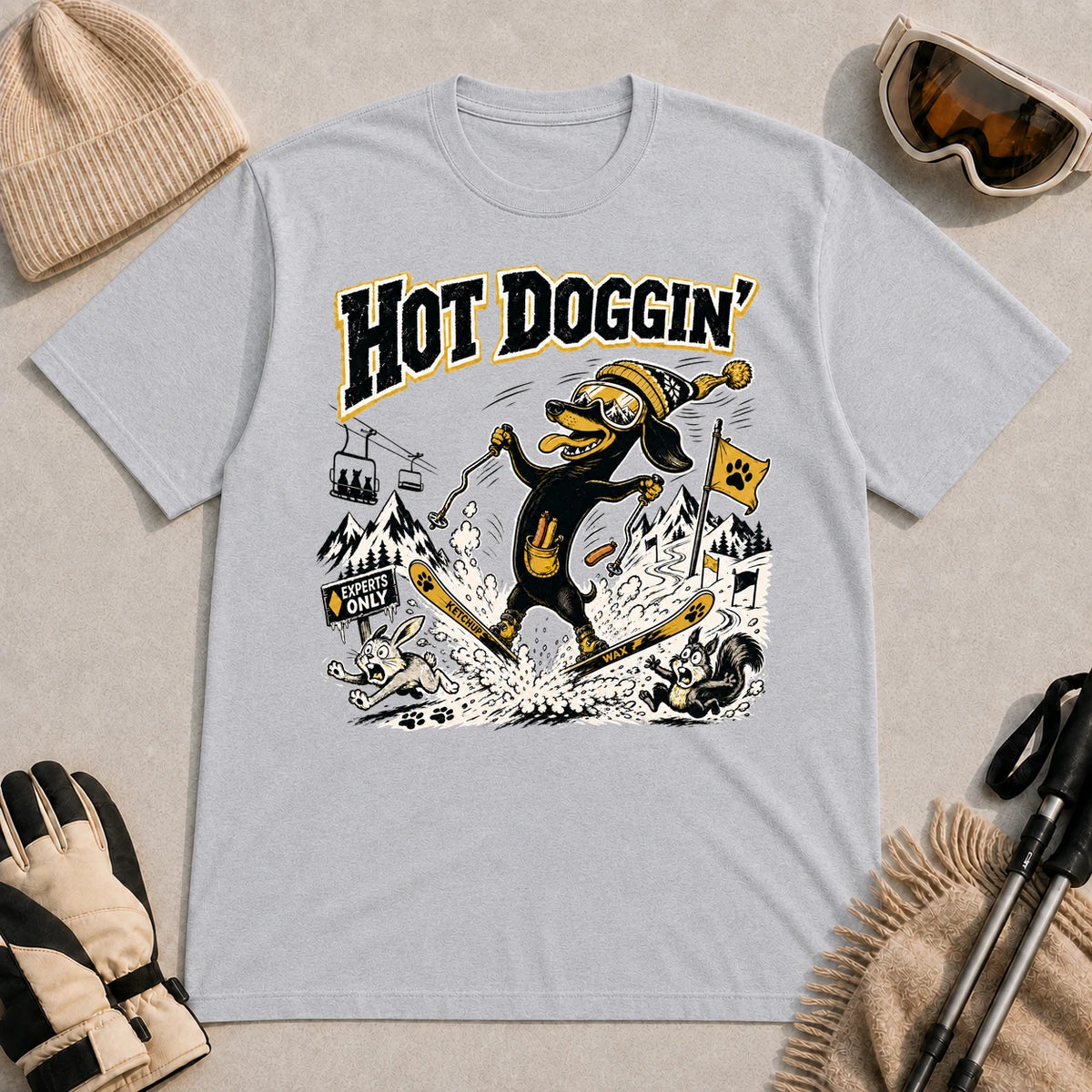 Sport Grey tee featuring a goggle-wearing dog skiing in a spread-eagle pose while startling woodland animals, with the phrase “Hot Doggin’.”