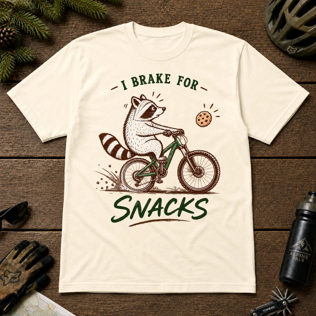 Natural tee featuring a raccoon mountain biking toward a cookie, with the phrase “I Brake for Snacks.”