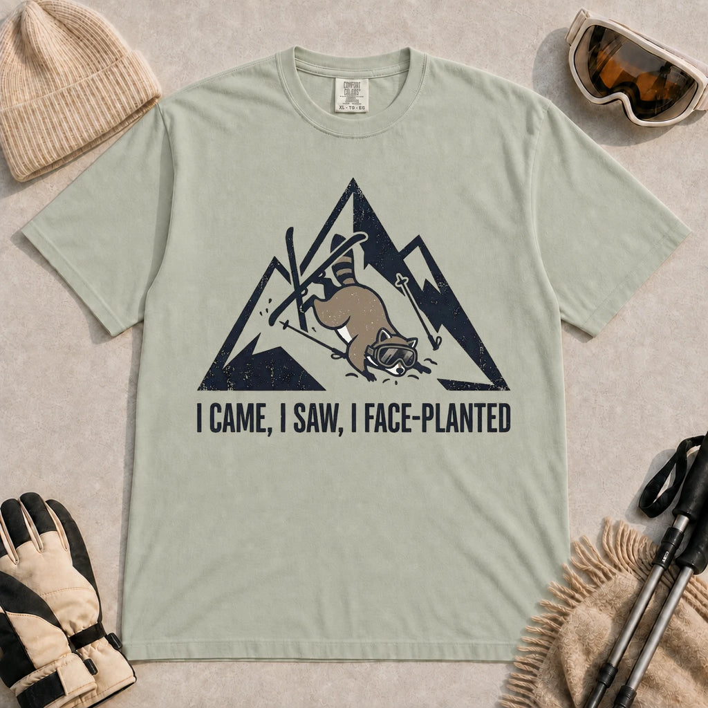 Bay tee featuring a raccoon face-planting into the snow beneath a distressed mountain graphic with the phrase “I Came, I Saw, I Face-Planted.”