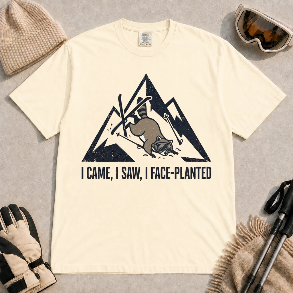 Ivory tee featuring a raccoon face-planting into the snow beneath a distressed mountain graphic with the phrase “I Came, I Saw, I Face-Planted.”