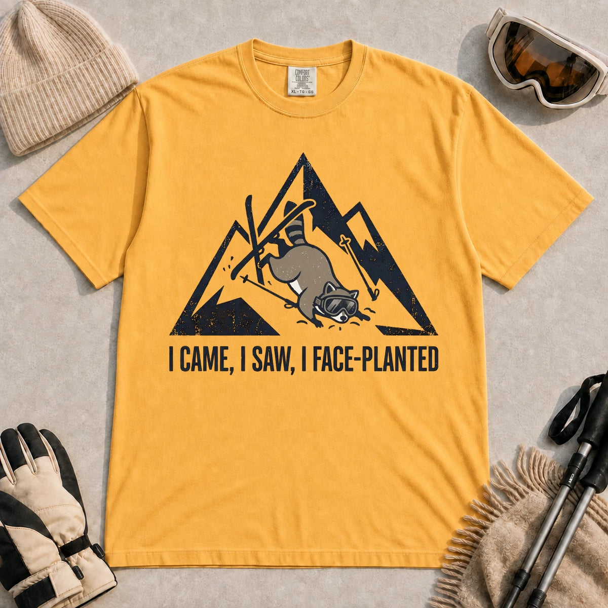 Mustard tee featuring a raccoon face-planting into the snow beneath a distressed mountain graphic with the phrase “I Came, I Saw, I Face-Planted.”