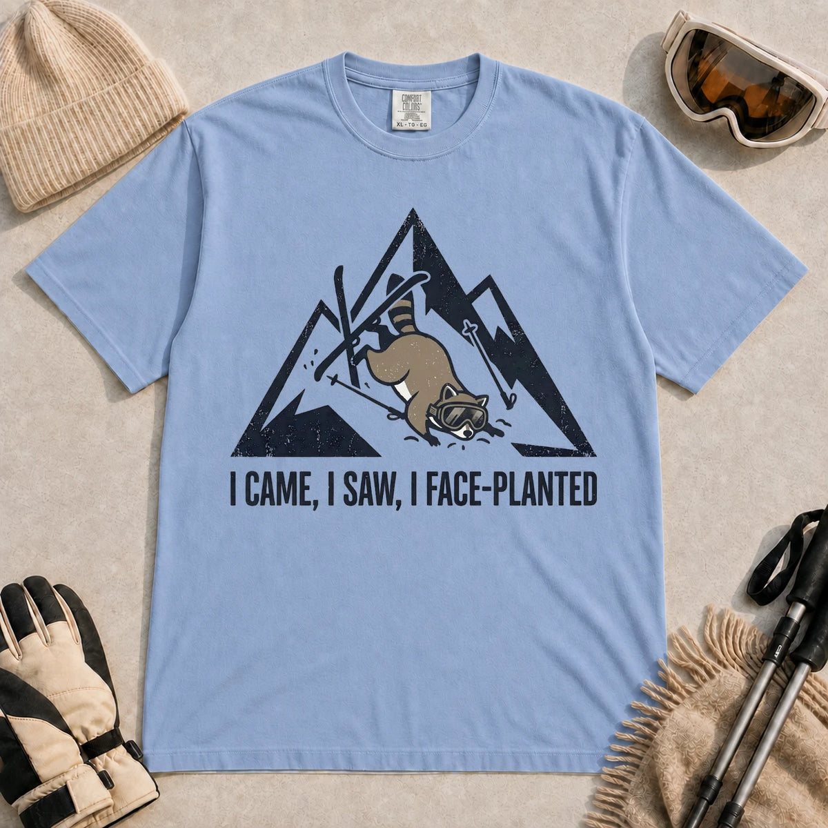 Washed Denim tee featuring a raccoon face-planting into the snow beneath a distressed mountain graphic with the phrase “I Came, I Saw, I Face-Planted.”