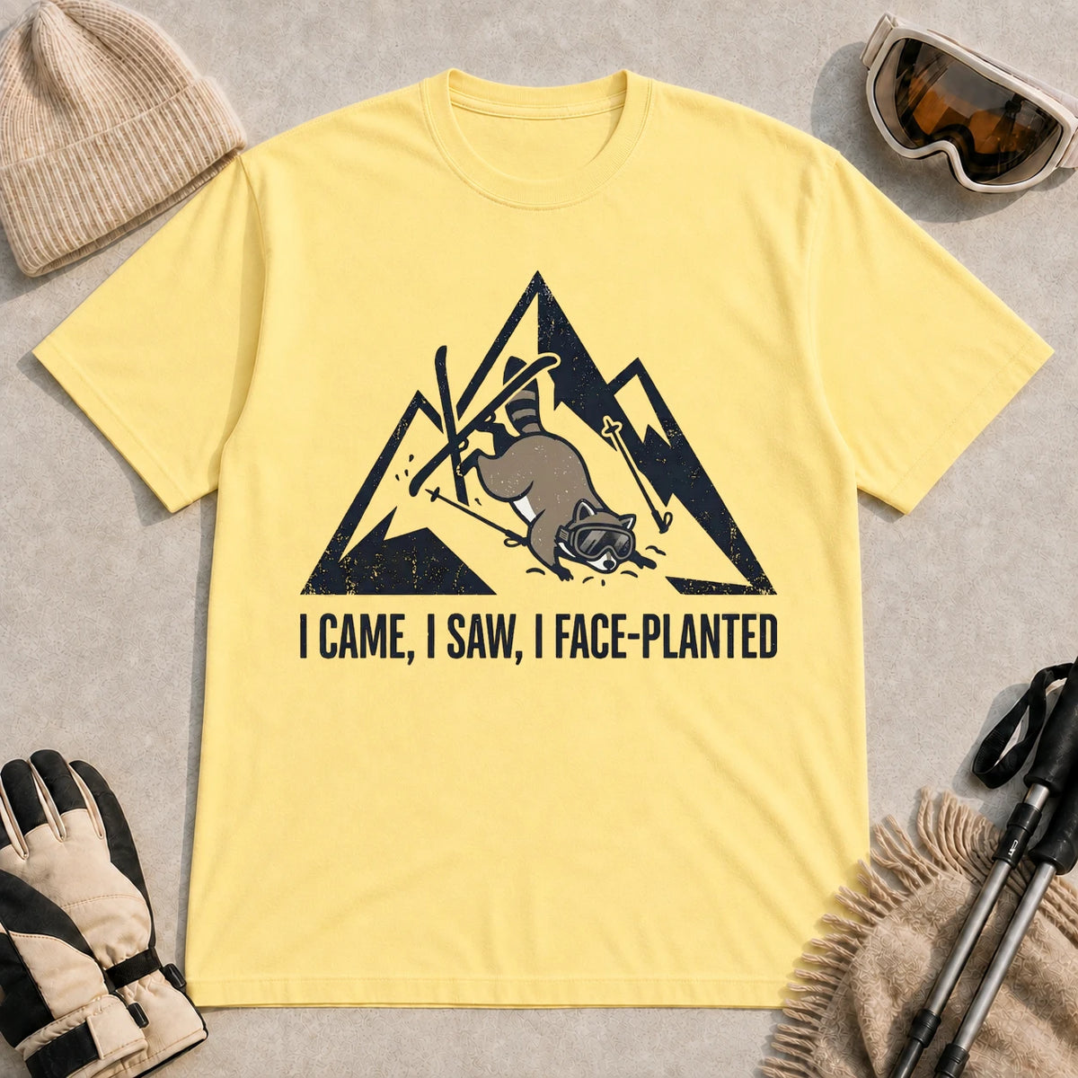 Cornsilk tee featuring a raccoon face-planting into the snow beneath a distressed mountain graphic with the phrase “I Came, I Saw, I Face-Planted.”