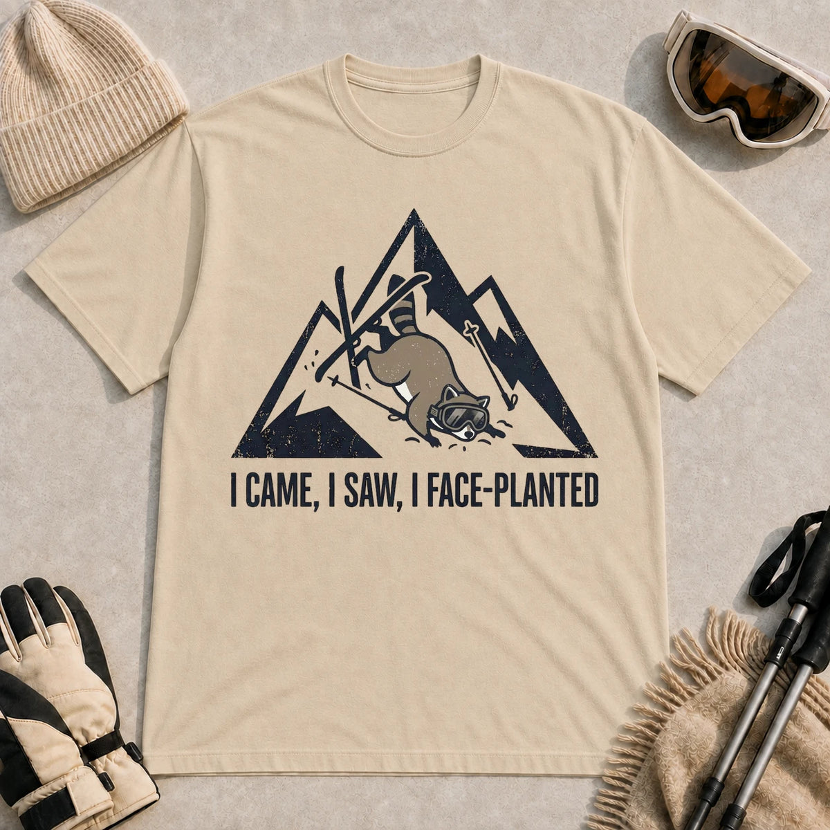 Sand tee featuring a raccoon face-planting into the snow beneath a distressed mountain graphic with the phrase “I Came, I Saw, I Face-Planted.”